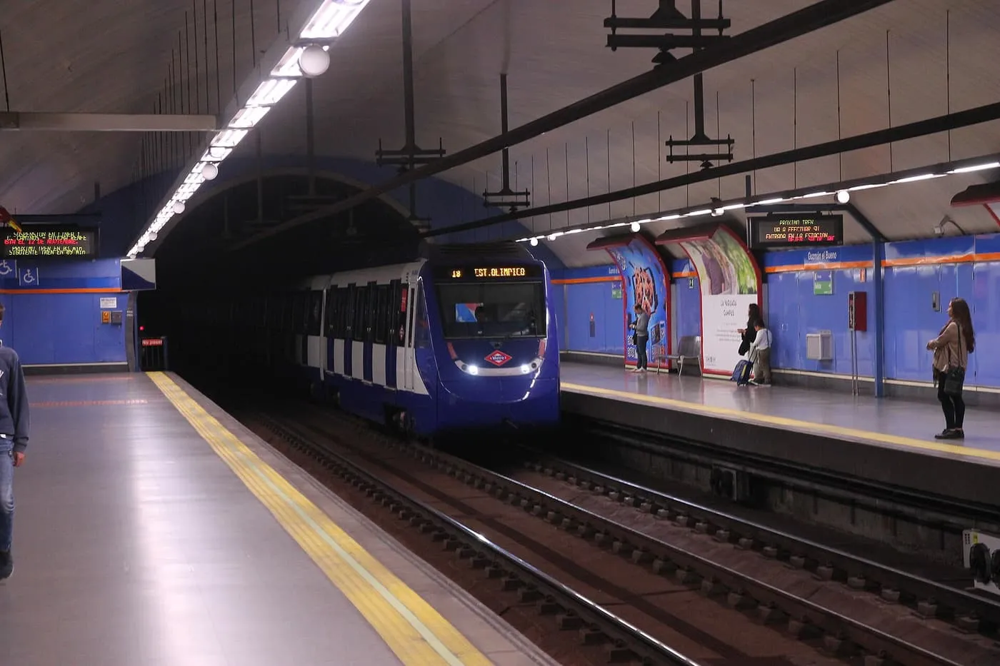 A blue and white Metro train arriving at the platform of Guzmán el Bueno station, under a low vaulted ceiling