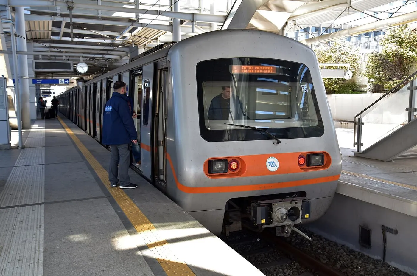 A grey and orange Athens Metro train standing at the platform of Athens International Airport station with its doors open