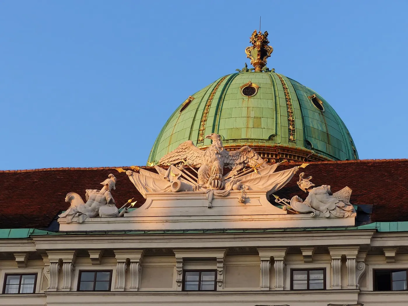 The green copper dome of the Michaelertor above the Hofburg's roofline, with a stone eagle and trophies on the pediment below it