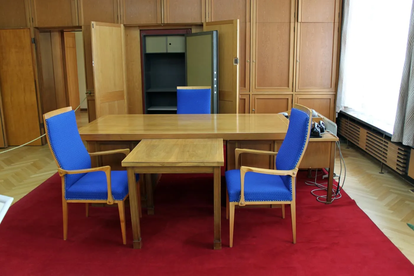 A wooden desk with two blue armchairs on a red carpet, and an open safe in the panelled wall behind it