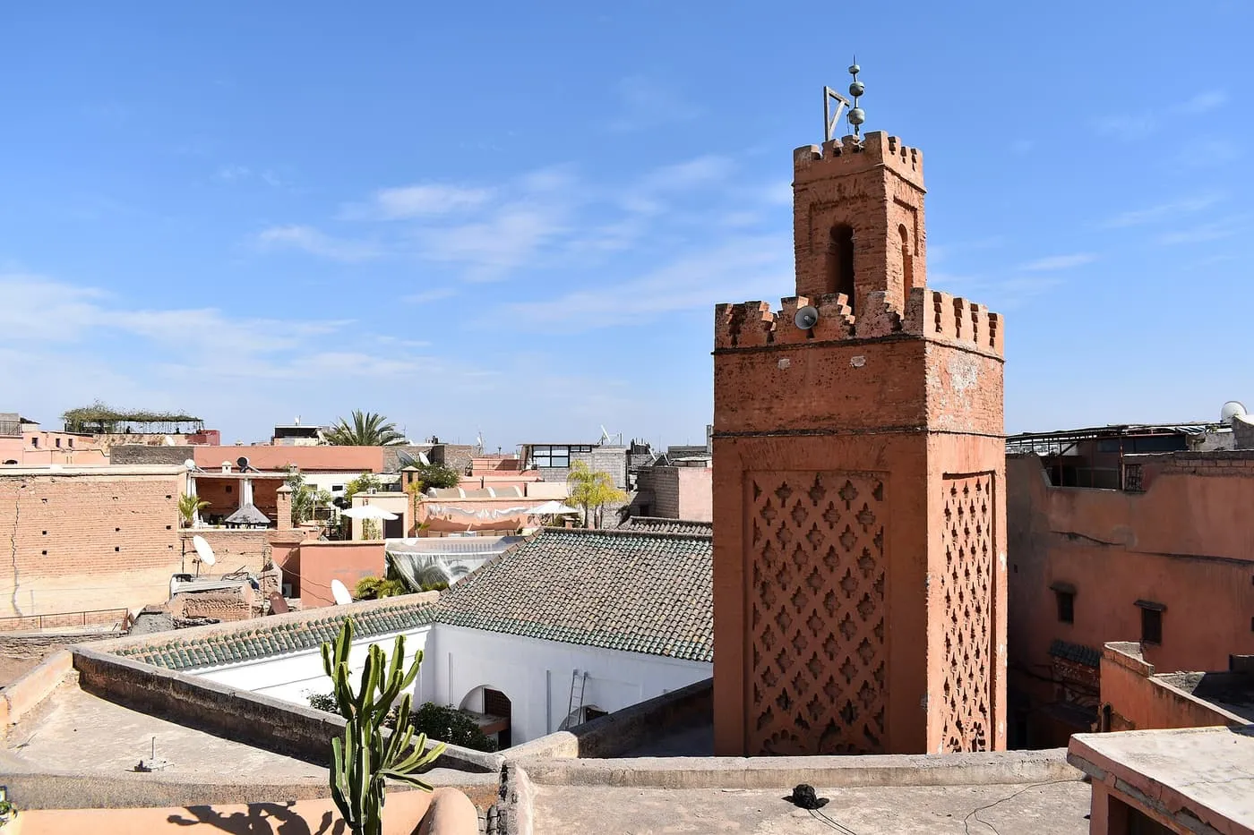 A brick minaret with lattice panels above flat rooftops and a courtyard in the Marrakech medina