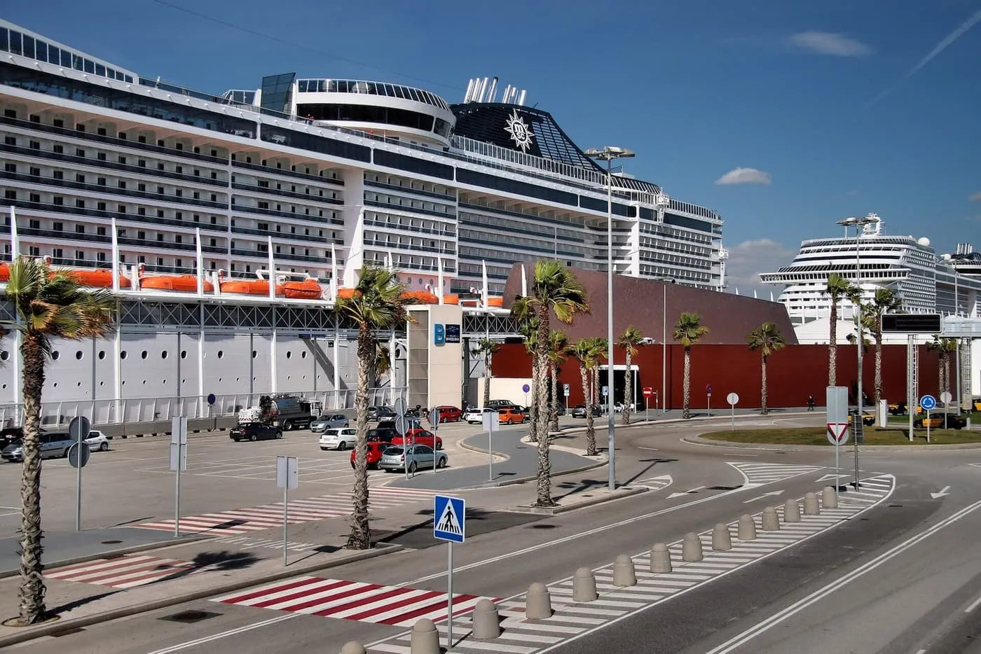 A cruise ship berthed beside a terminal building and car park on the Moll Adossat, with palm trees along the road