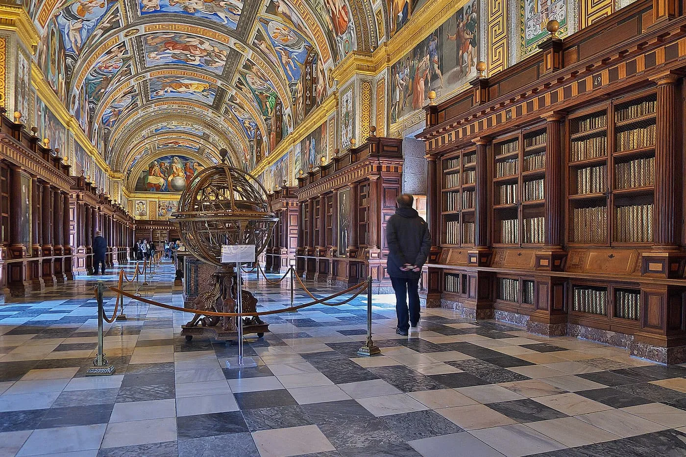 The barrel-vaulted Library of El Escorial with frescoed ceiling, dark wooden bookcases and a bronze armillary sphere on a chequered marble floor