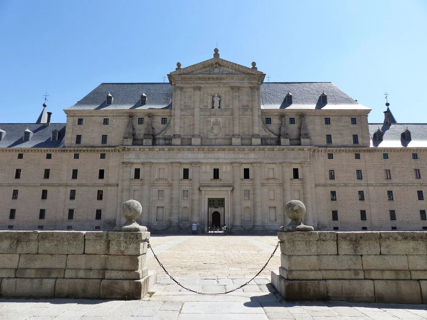 The west façade of the monastery of El Escorial, a long granite front with a central pediment and slate roof, seen across a stone wall
