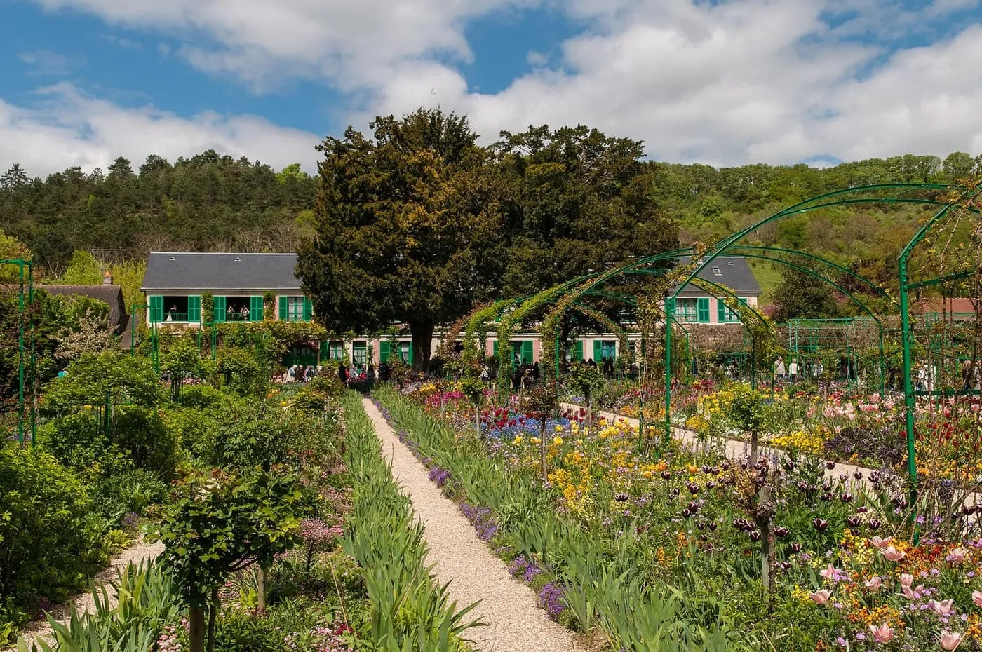 Monet's house at the end of a gravel path between beds of spring flowers, with green metal arches over the path