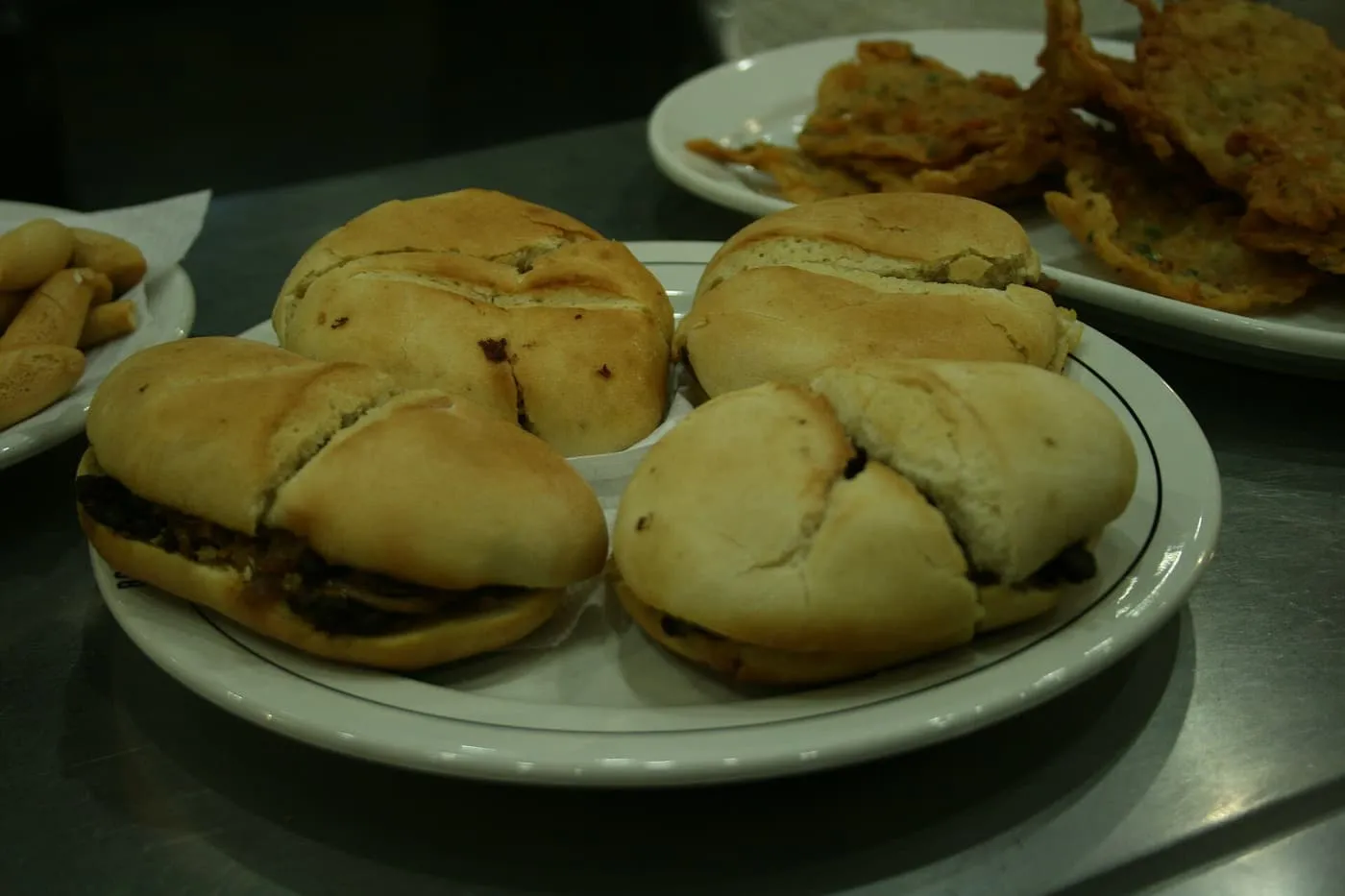 Three small bread rolls on a plate, each filled with dark stewed meat, beside a plate of fried food