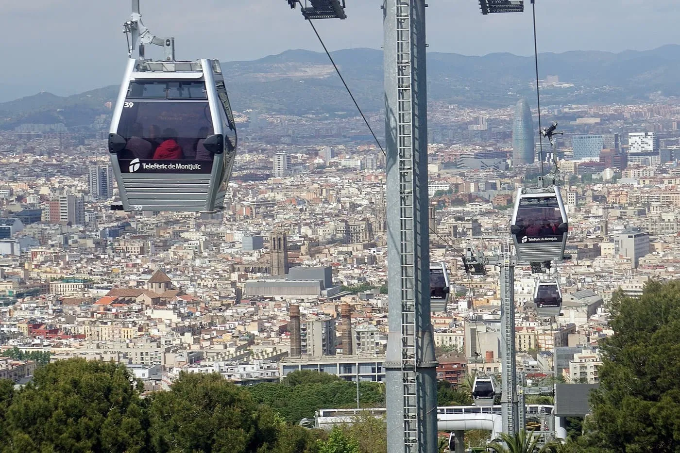 Grey Montjuïc cable-car cabins on their wires above Barcelona's rooftops, with the Torre Glòries and hills behind