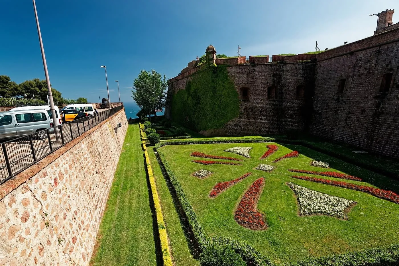The brick walls of Montjuïc Castle above a clipped lawn planted with a red and white flower pattern, in the old moat