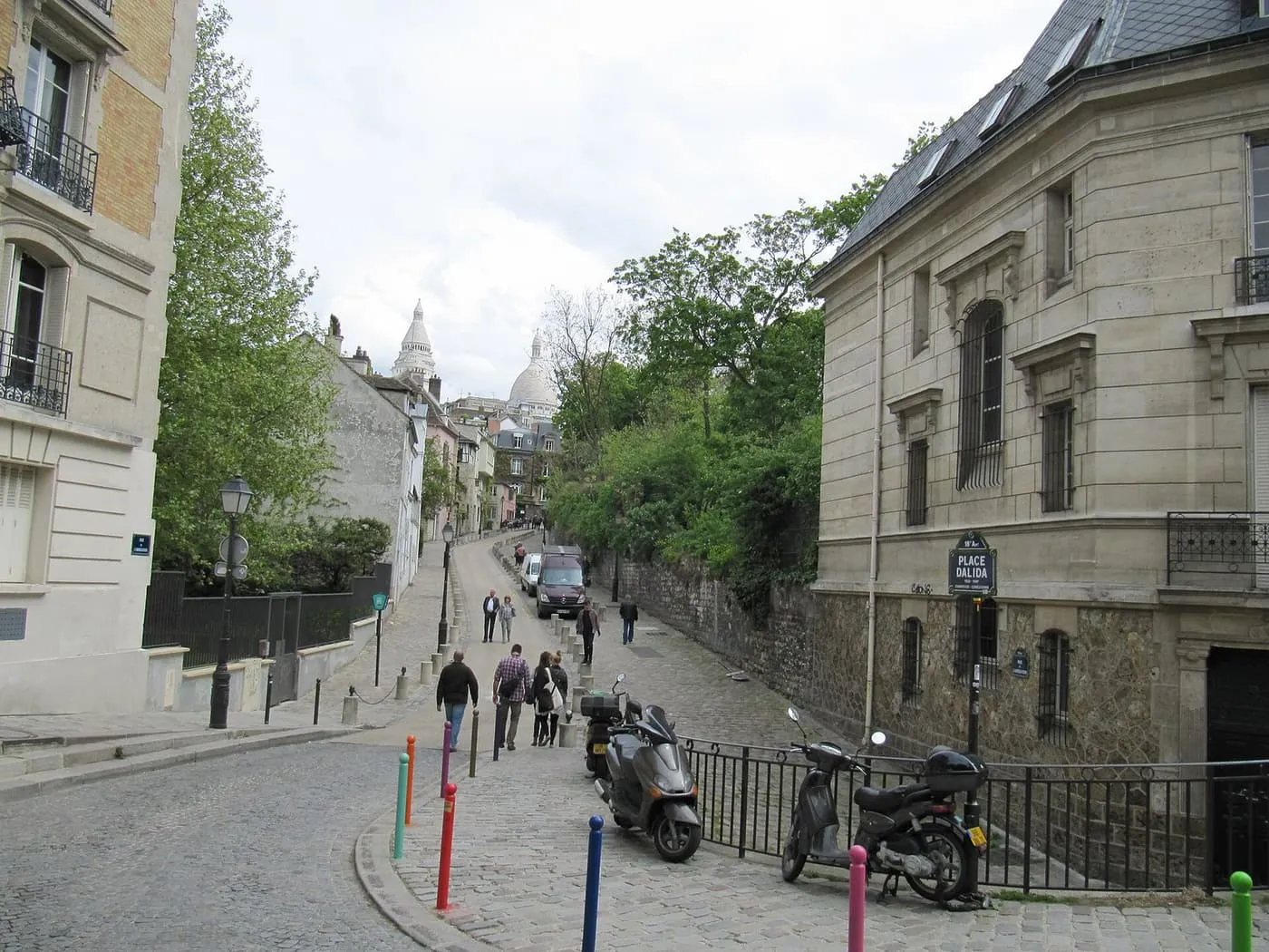 A cobbled street climbing through Montmartre from Place Dalida, with the white domes of Sacré-Cœur at the top