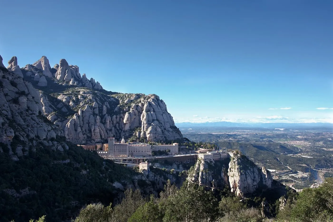 The monastery of Montserrat on a ledge below grey rock pinnacles, with the plain stretching away to distant mountains