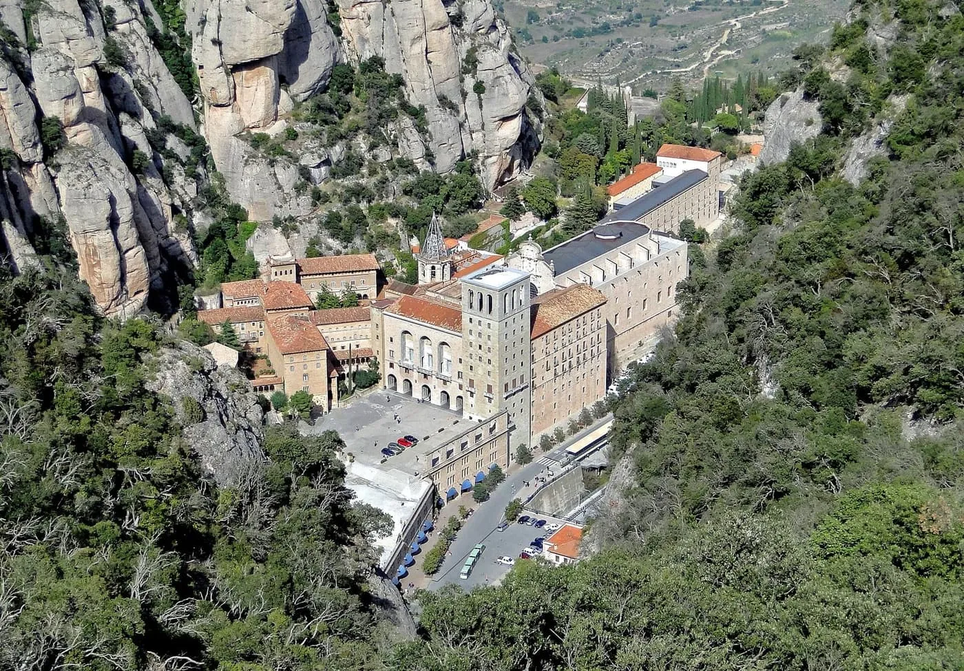 The monastery of Montserrat seen from above, its basilica, bell tower and square set on a ledge below grey rock pinnacles