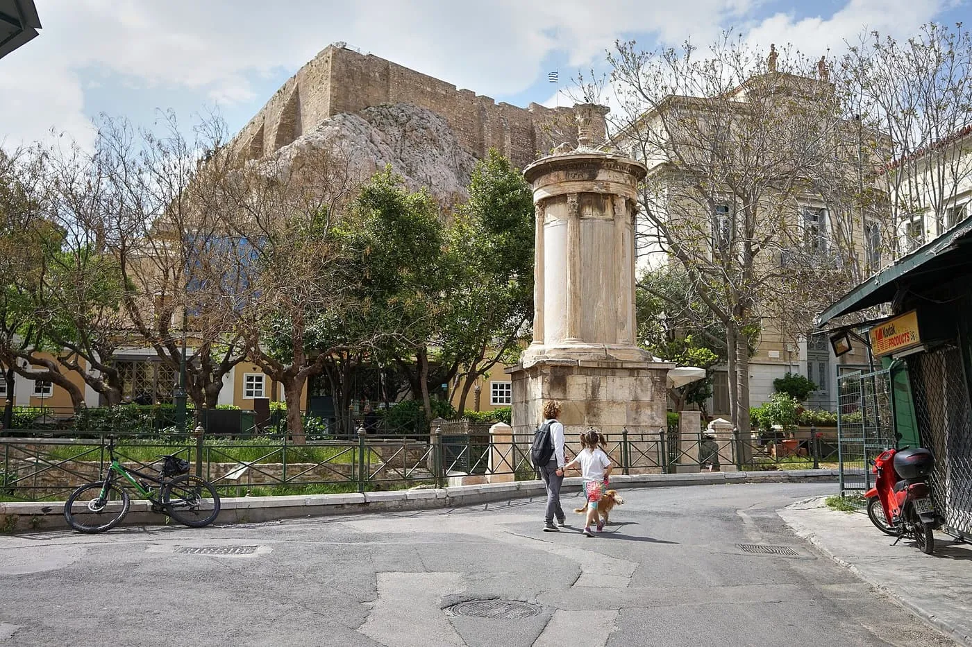 The Choragic Monument of Lysicrates, a round marble tower on a square base, with the Acropolis wall above the trees behind it