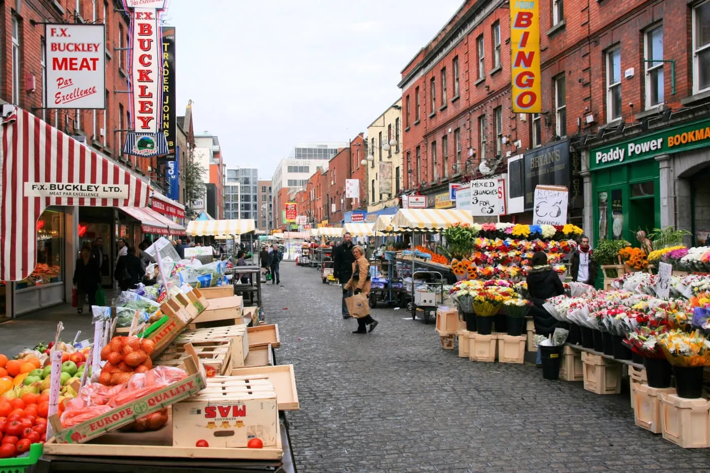 Fruit and flower stalls and striped awnings on the cobbled length of Moore Street market, with brick buildings either side