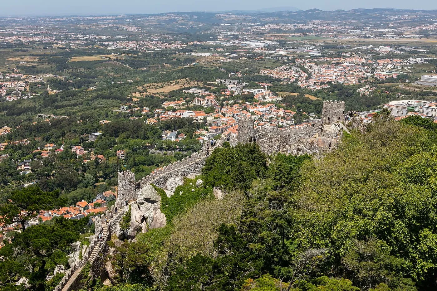 Granite walls of the Moorish Castle running along a wooded ridge, with Sintra's rooftops and the plain below