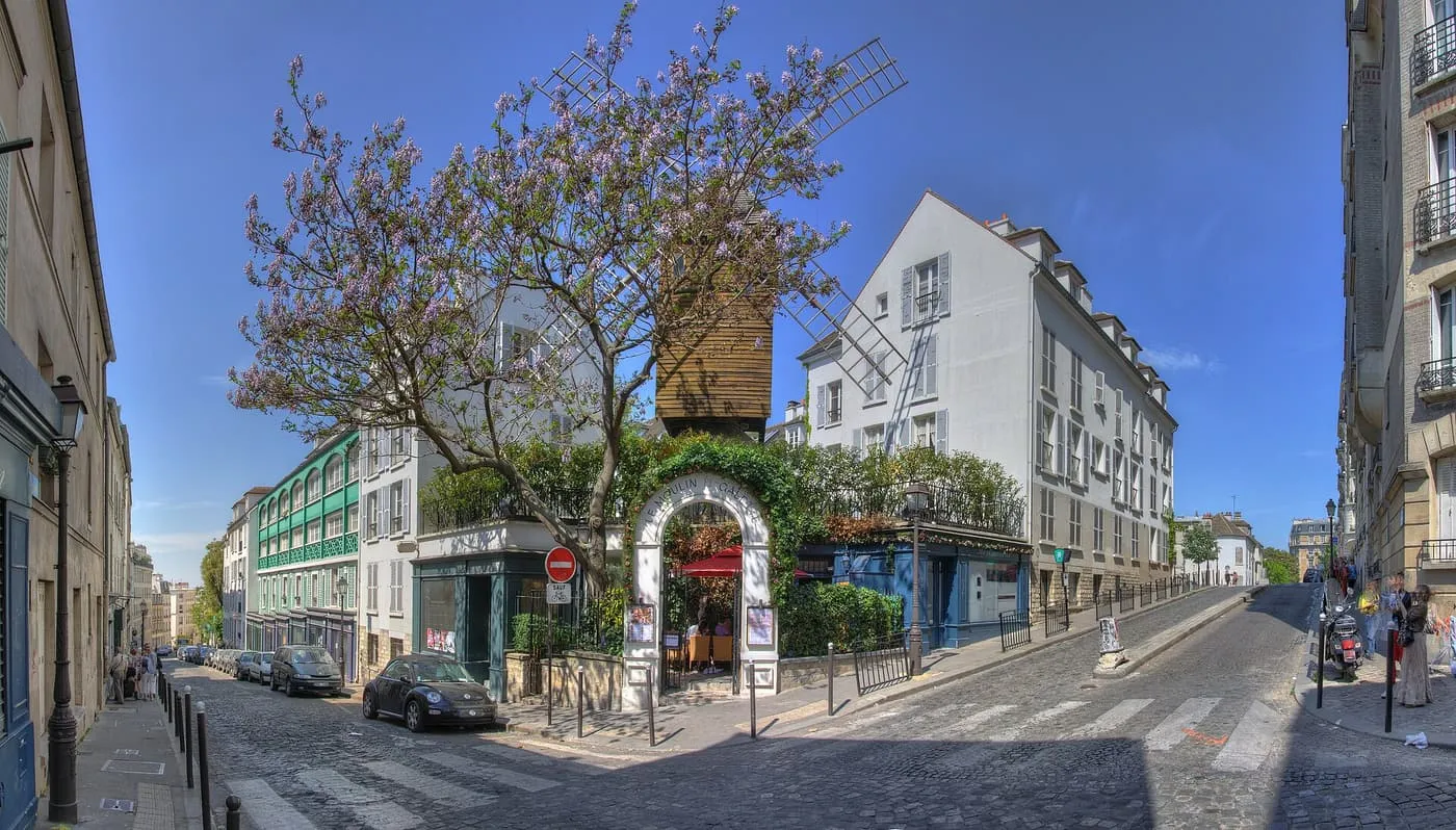 The wooden Moulin de la Galette windmill rising behind a vine-covered gateway on the corner of two cobbled streets, under a flowering tree
