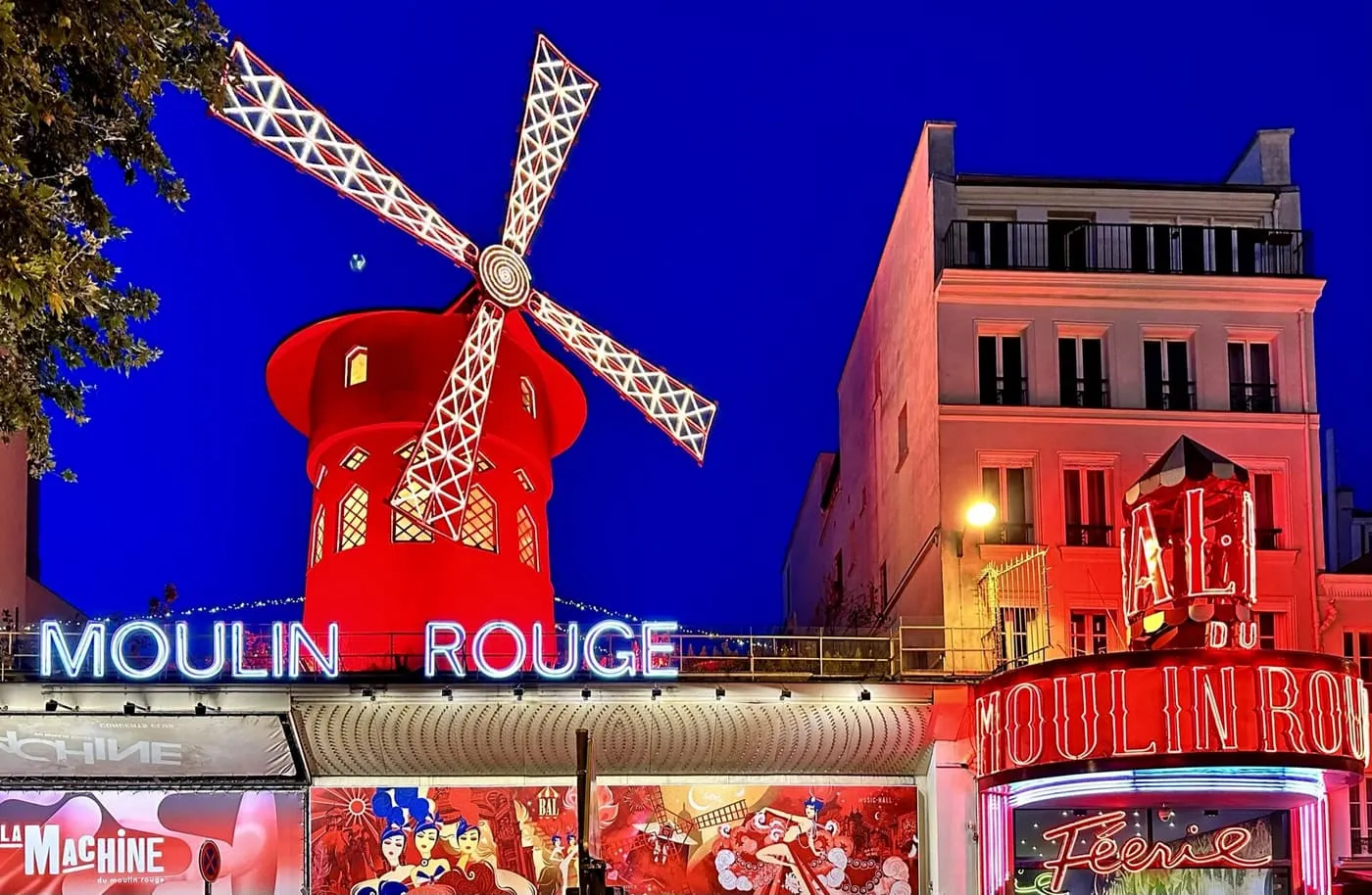 The Moulin Rouge at night, a red windmill with white-lit sails above its blue and red neon sign, and the Féerie sign on the entrance