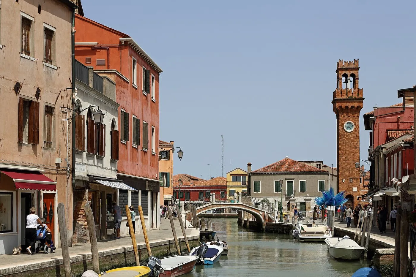 A canal on Murano lined with shops and houses, with a stone bridge and the brick clock tower of Santo Stefano at the far end