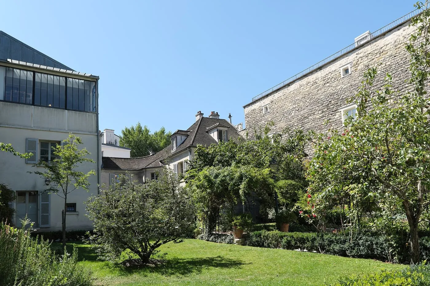 The Renoir gardens at the Musée de Montmartre, lawn and shrubs in front of pale buildings and a tall stone wall under a blue sky