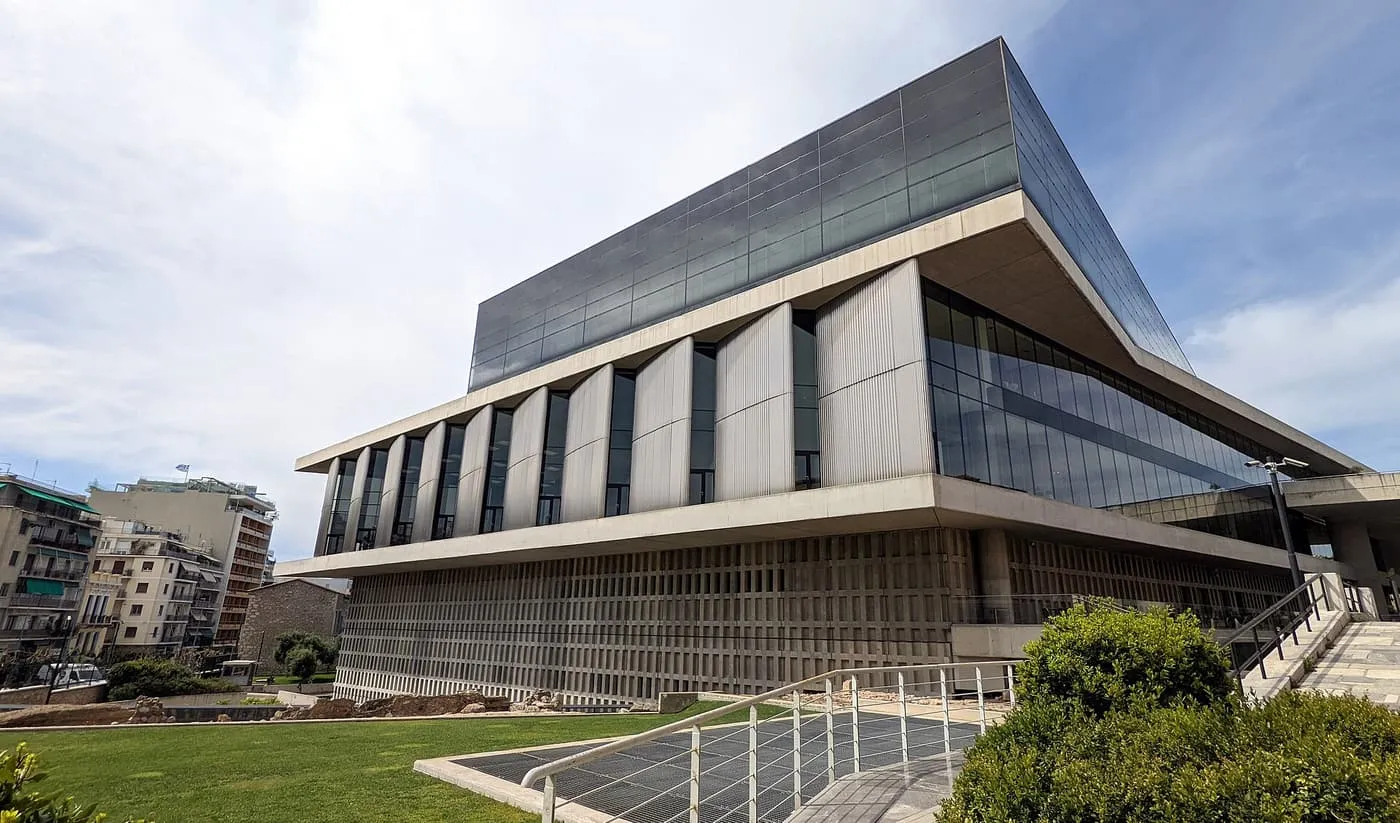 The Acropolis Museum, a dark glass block above angled concrete floors, with a lawn and a metal grille walkway in front