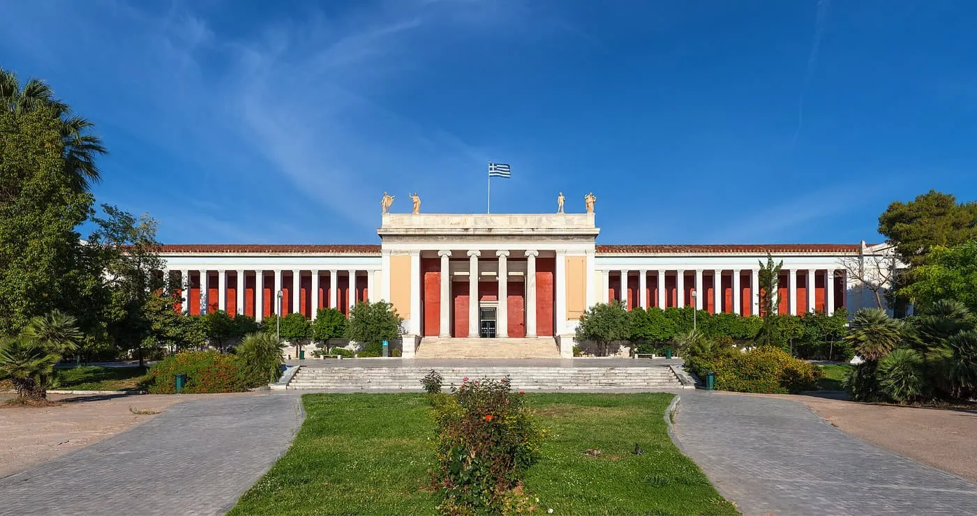The neoclassical front of the National Archaeological Museum, a white portico with red walls and statues on the roof, behind a lawn and two paths