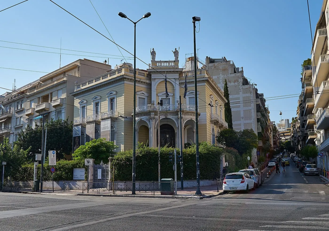 The yellow neoclassical Museum of Cycladic Art on a street corner, with a rounded portico and statues on the roofline