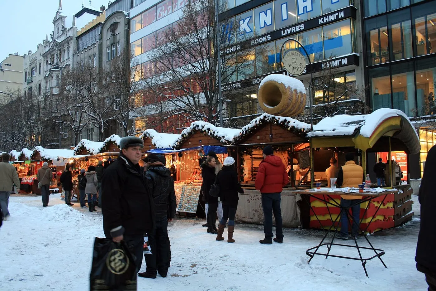 Wooden Christmas market huts with snow on their roofs at Můstek, at the lower end of Wenceslas Square, with a giant model chimney cake over one stall and shoppers in winter coats