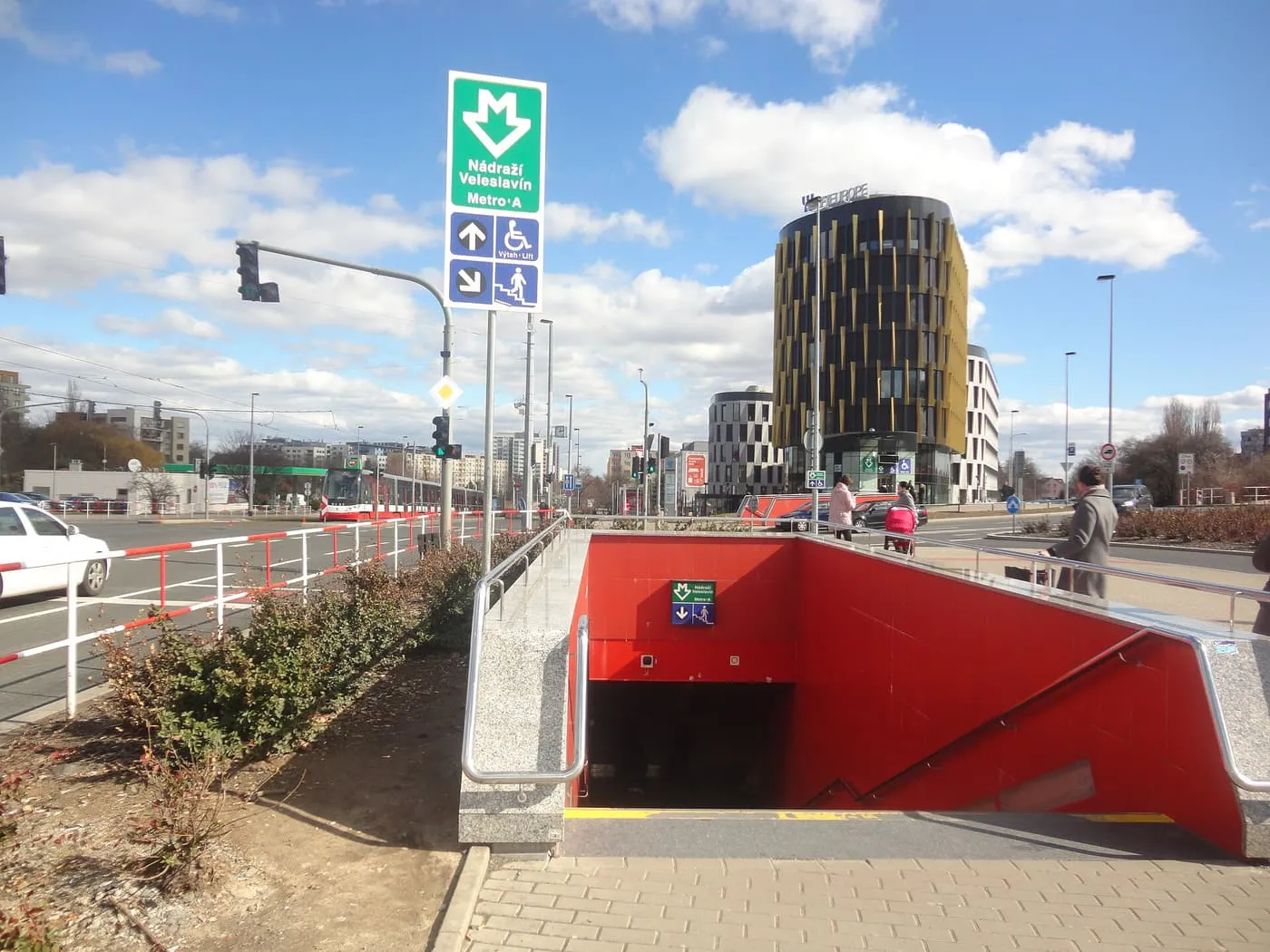 The red-tiled entrance stairs to Nádraží Veleslavín metro station, beside a green Metro A sign, with an office tower behind