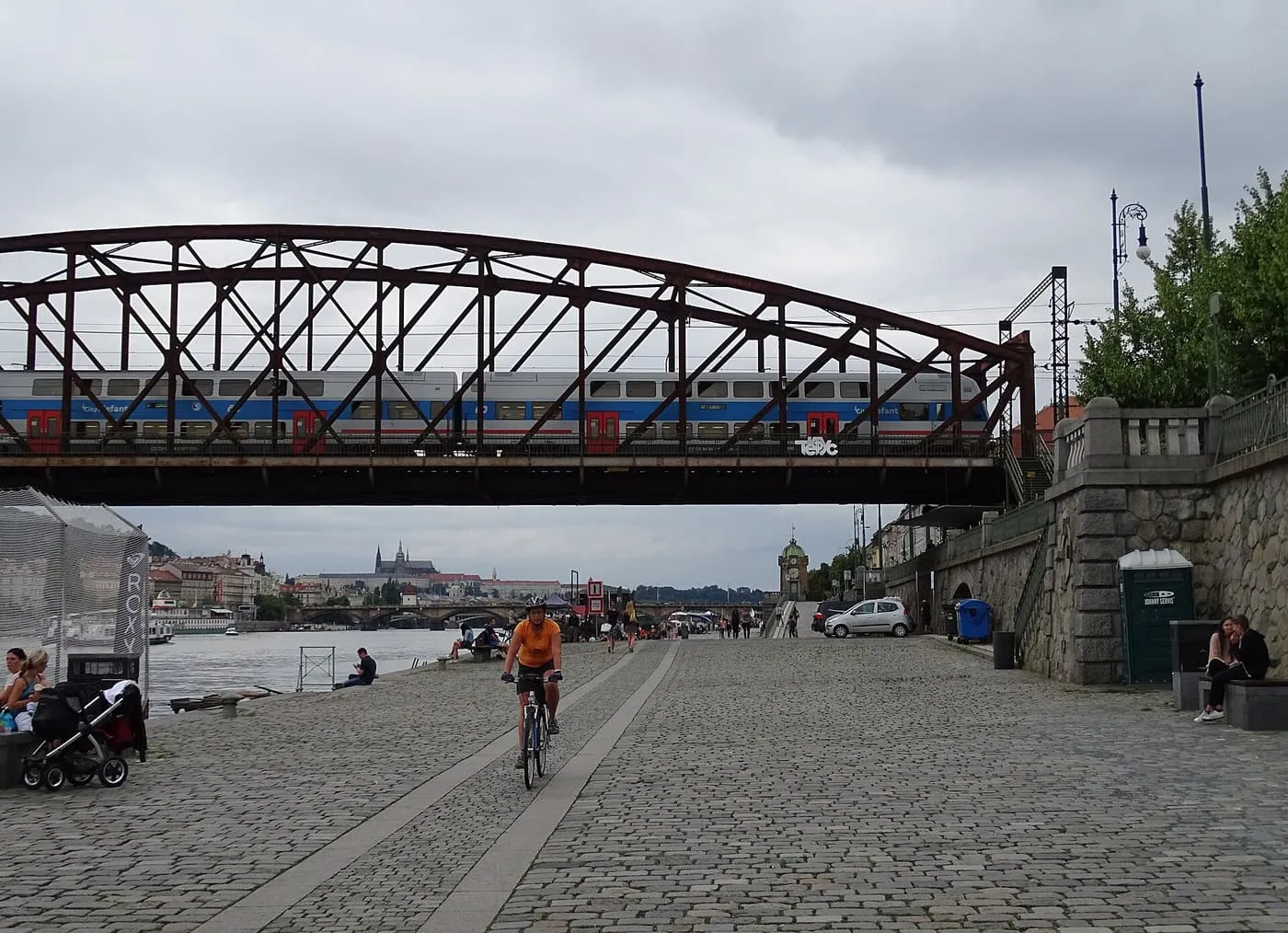 The cobbled Rašín Embankment at Náplavka, with a cyclist passing under a steel railway bridge carrying a train, and Prague Castle on the skyline in the distance