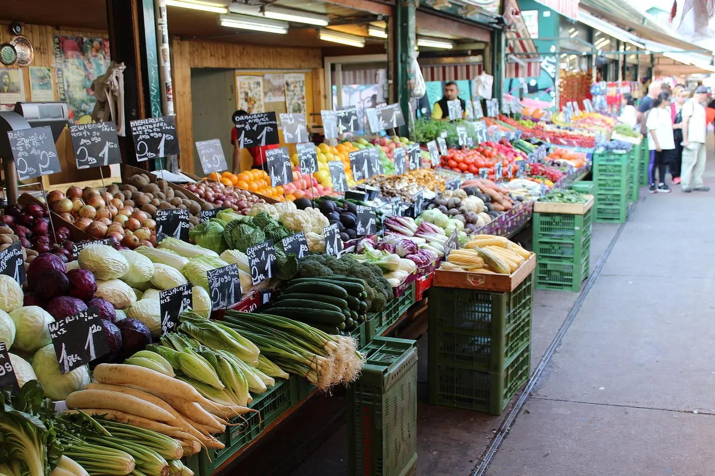 Naschmarkt stalls piled with cabbages, leeks, radishes and other vegetables under chalkboard price tags, with shoppers further along the aisle