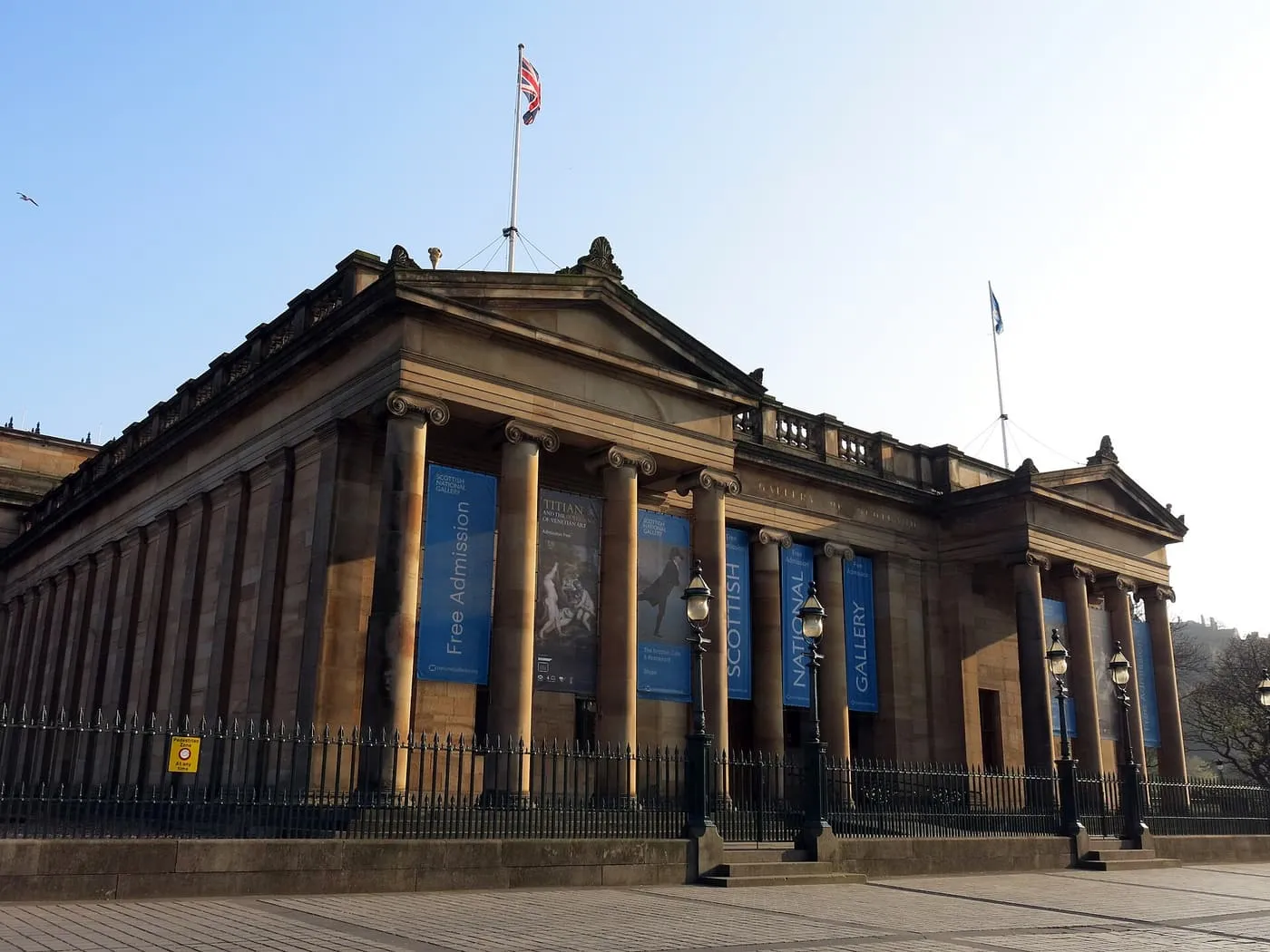 The columned, pedimented stone front of the National Gallery on the Mound, with flags on the roof and exhibition banners between the columns