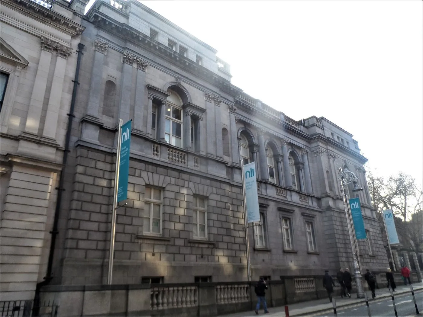 The grey stone side of the National Library of Ireland on Kildare Street, with blue banners on lamp posts along the pavement