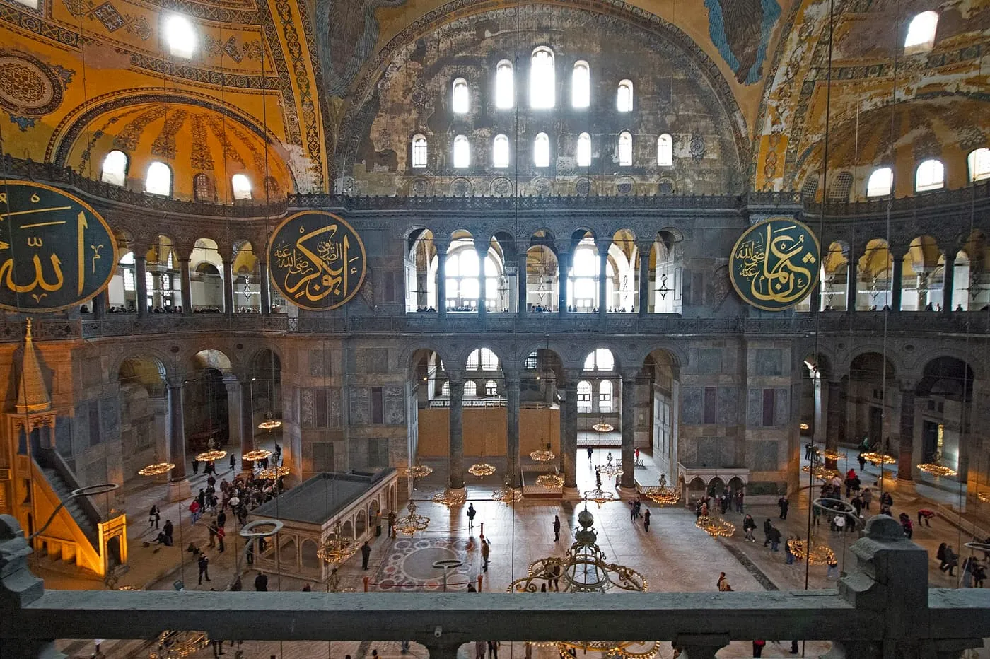 The nave of Hagia Sophia seen over the gallery balustrade, with large calligraphic roundels, chandeliers and visitors on the floor far below