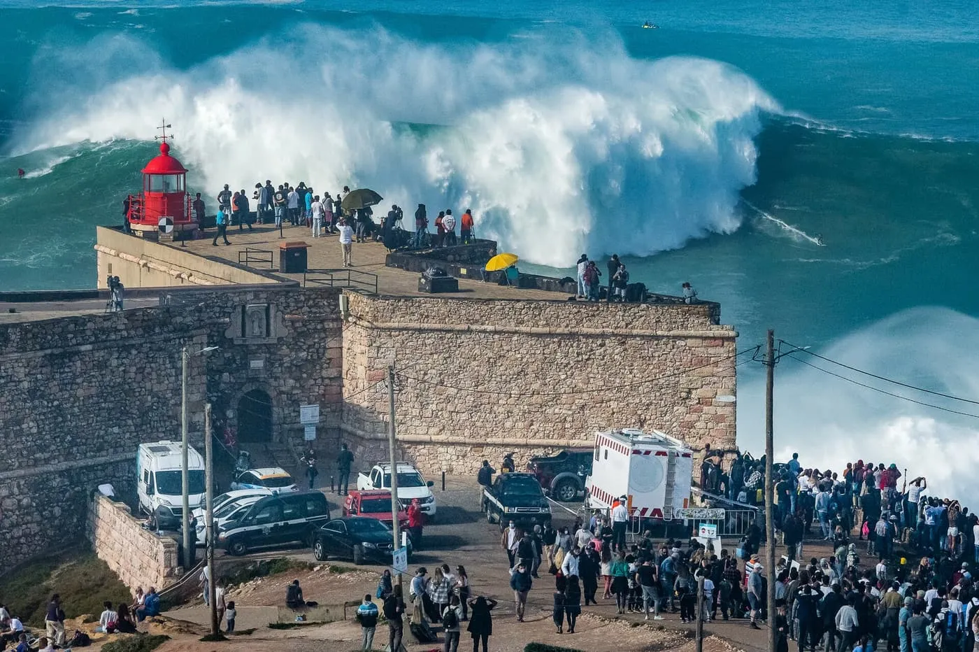 A huge wave breaking behind the red lighthouse and fort at Nazaré, with crowds watching from the clifftop