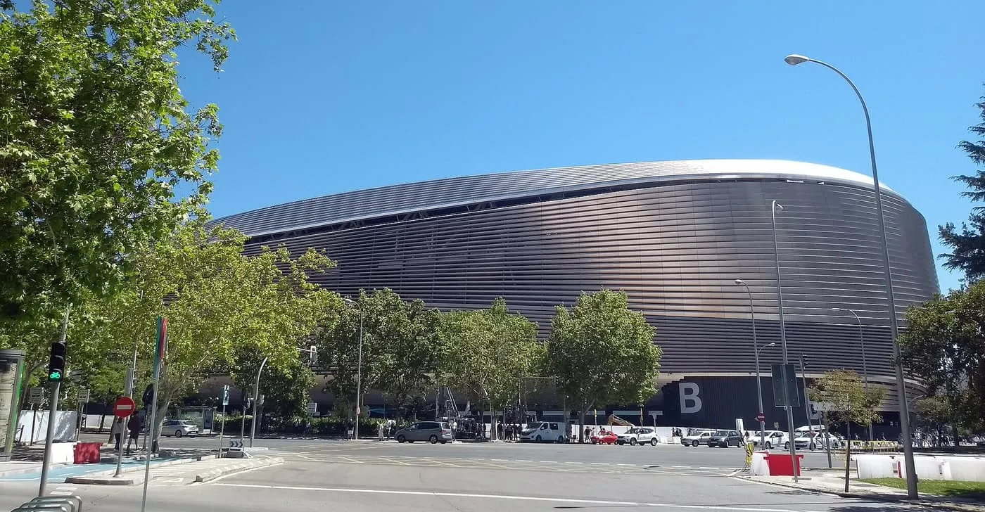 The curved west façade of the rebuilt Santiago Bernabéu, clad in horizontal steel slats under a blue sky, seen across a street