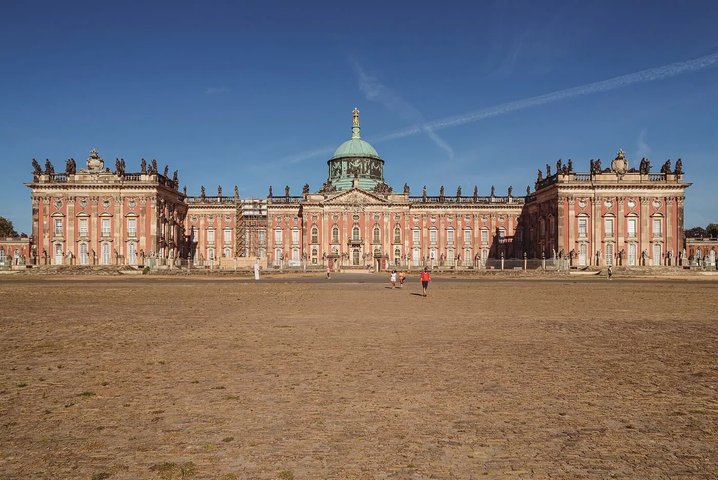 The long pink and sandstone front of the New Palace in Potsdam, with a green copper dome, seen across its forecourt