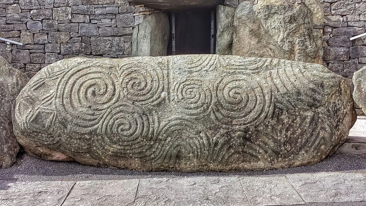 A large boulder carved with spirals and lozenges in front of the dark entrance to the Newgrange passage
