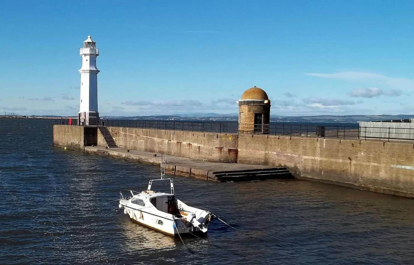 A white lighthouse and a small domed stone lookout on the harbour wall at Newhaven, with a motorboat moored in the foreground and the Firth of Forth beyond