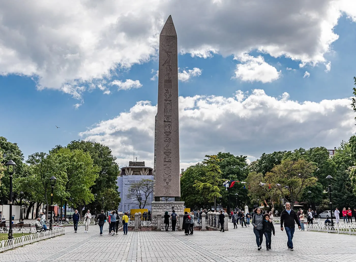 The Obelisk of Theodosius rising above trees and a paved square under a blue and white sky, with people walking across it