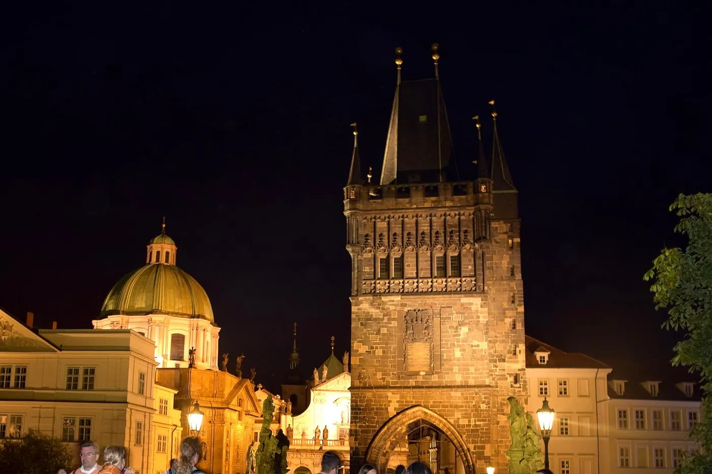 The Old Town Bridge Tower floodlit at night, a dark Gothic gate with a pointed roof, beside a green copper dome