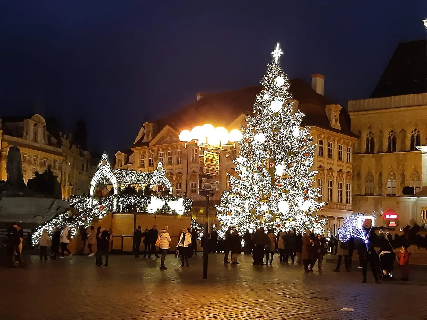 The lit Christmas tree and market stalls on Old Town Square in Prague at night, with people crossing the cobbles