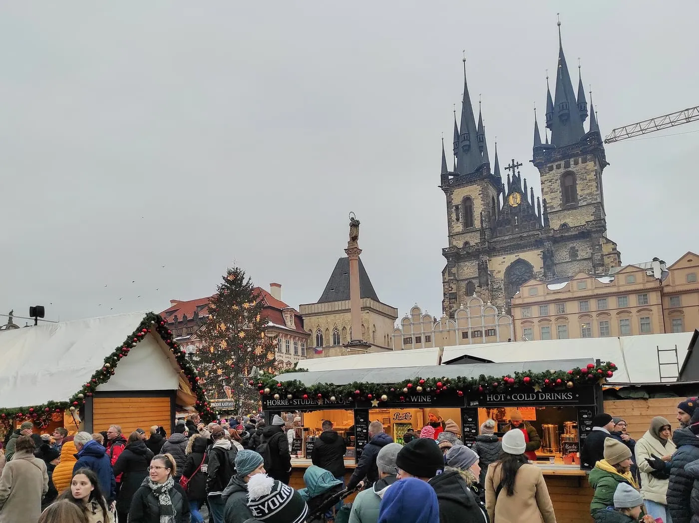 Wooden market stalls hung with garlands on Old Town Square in daylight, with the Christmas tree, the Marian column and the dark spires of Týn Church behind