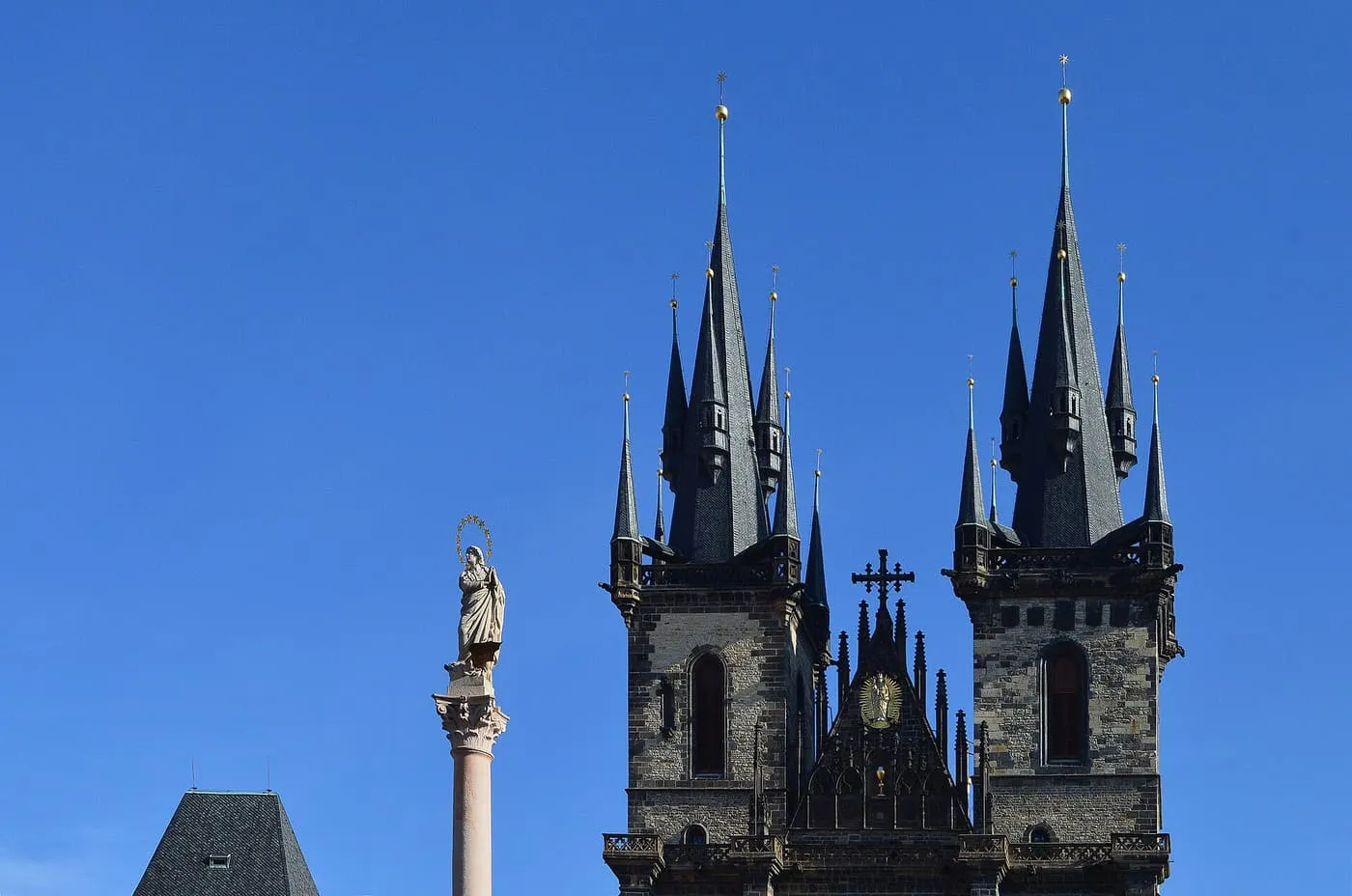 The twin Gothic spires of the Church of Our Lady before Týn against a clear blue sky, with a stone column topped by a statue to their left
