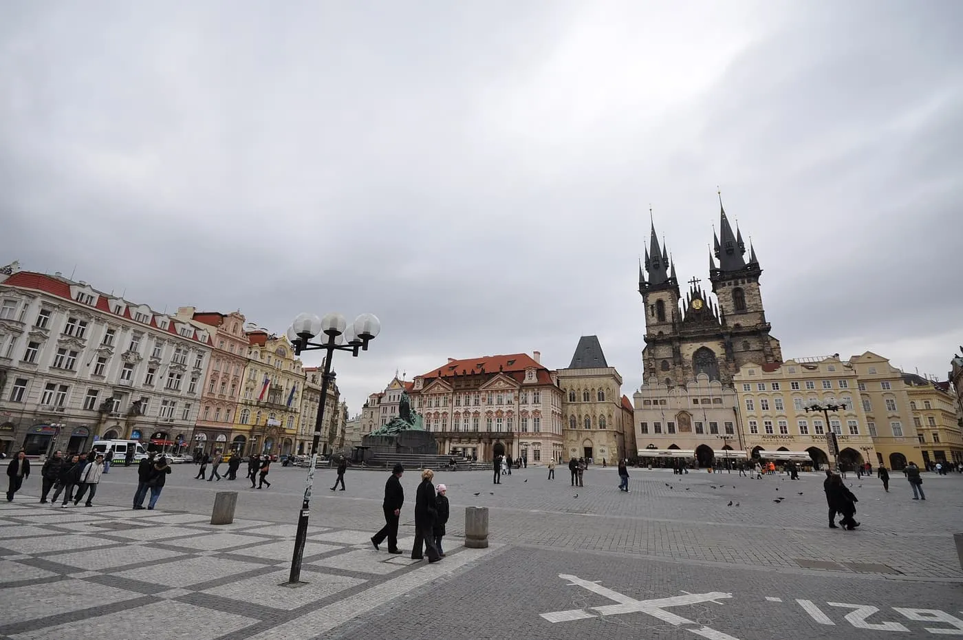 Old Town Square, a wide paved square with the twin towers of the Týn church and pastel townhouses on its far side under a grey sky