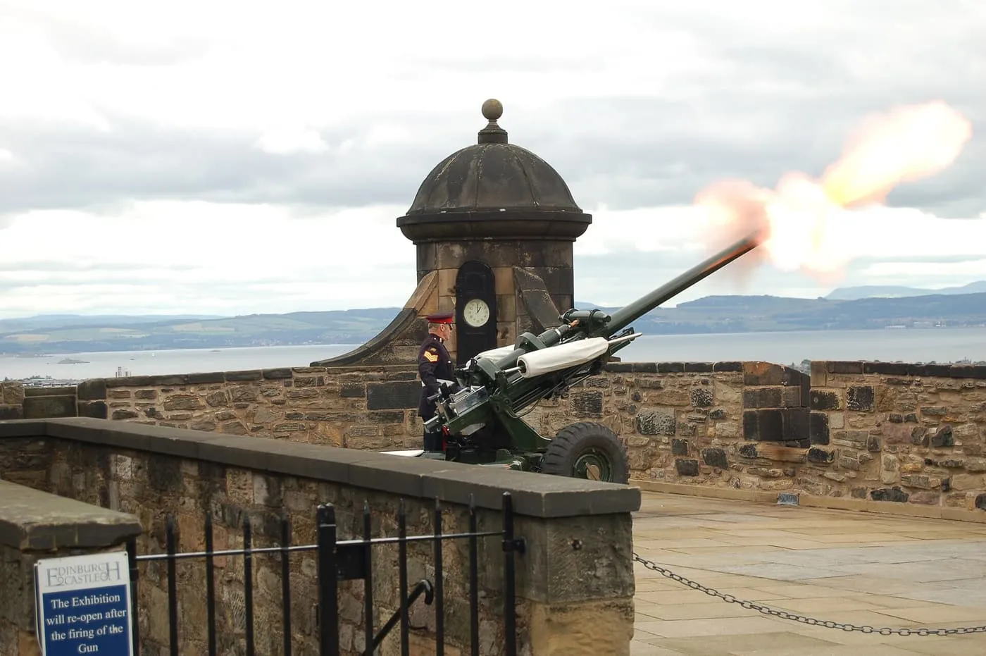 A soldier beside a field gun as it fires a flash of flame, with a small clock turret and the Firth of Forth behind