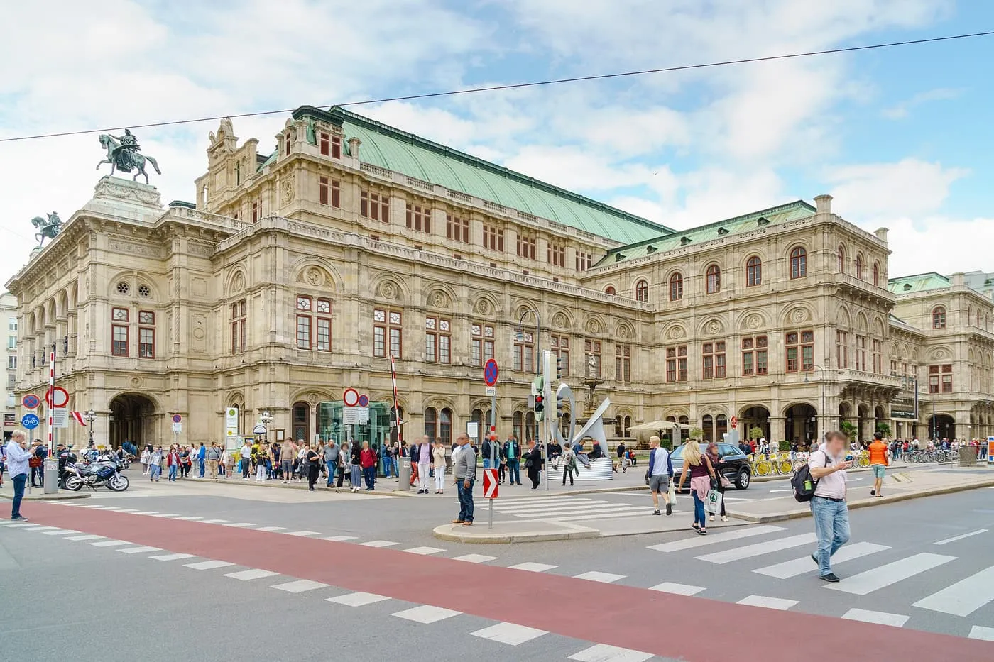 The Vienna State Opera, a Neo-Renaissance building with a green copper roof and equestrian statues on the corners, seen across the Ring with pedestrians at the crossing