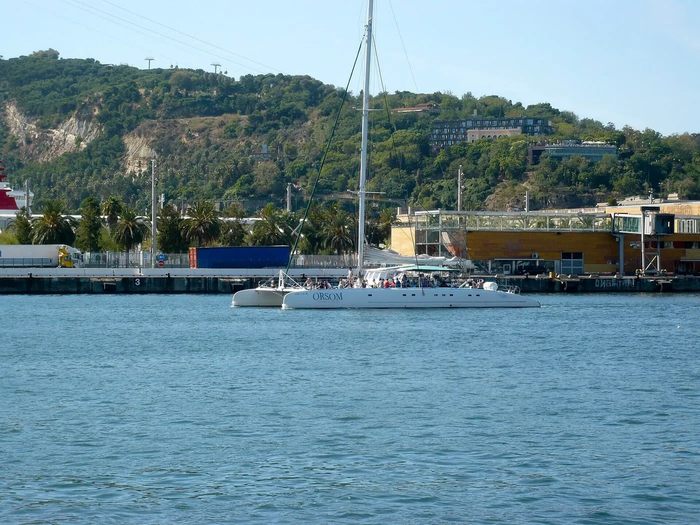 The white sailing catamaran Orsom crossing Barcelona harbour, with the wooded slope of Montjuïc behind