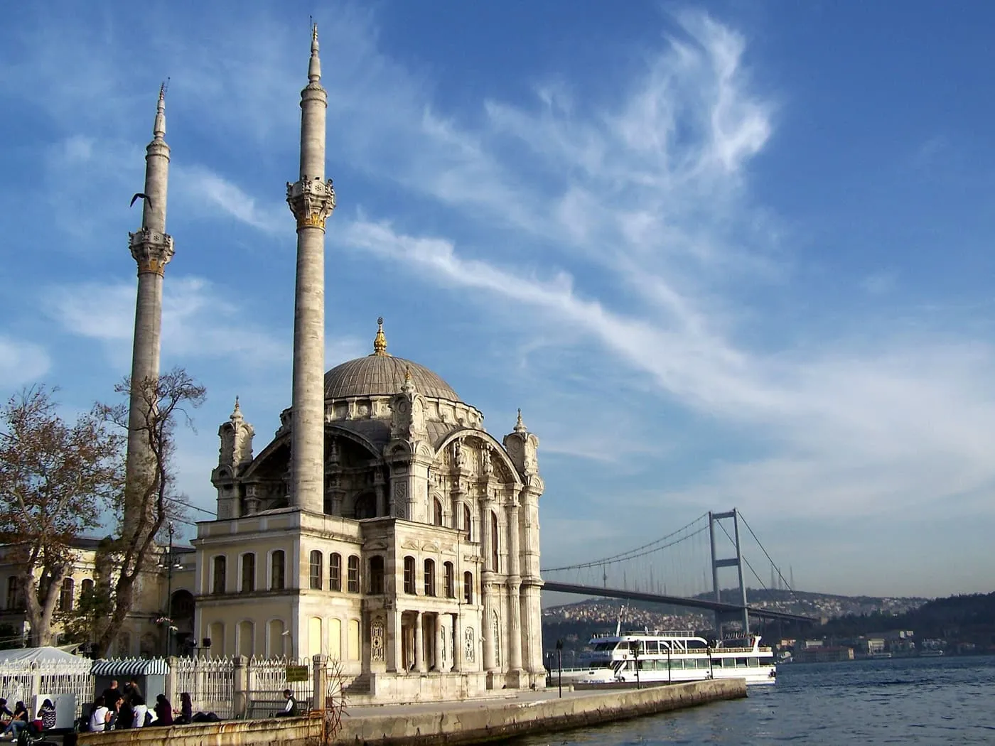 Ortaköy Mosque on the Bosphorus shore with its two slender minarets, the Bosphorus Bridge behind it and a white tour boat on the water