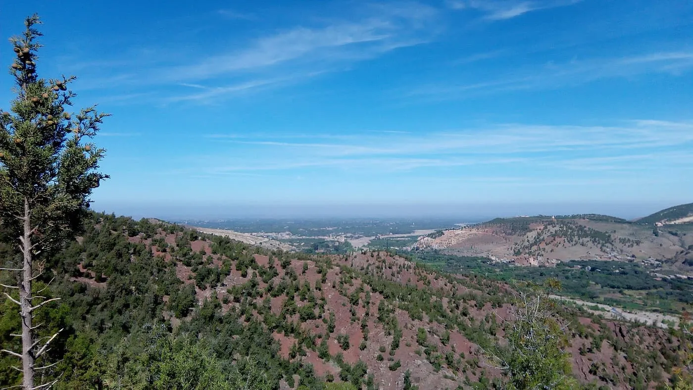 Red hillsides dotted with trees above the Ourika valley, with the plain hazy in the distance