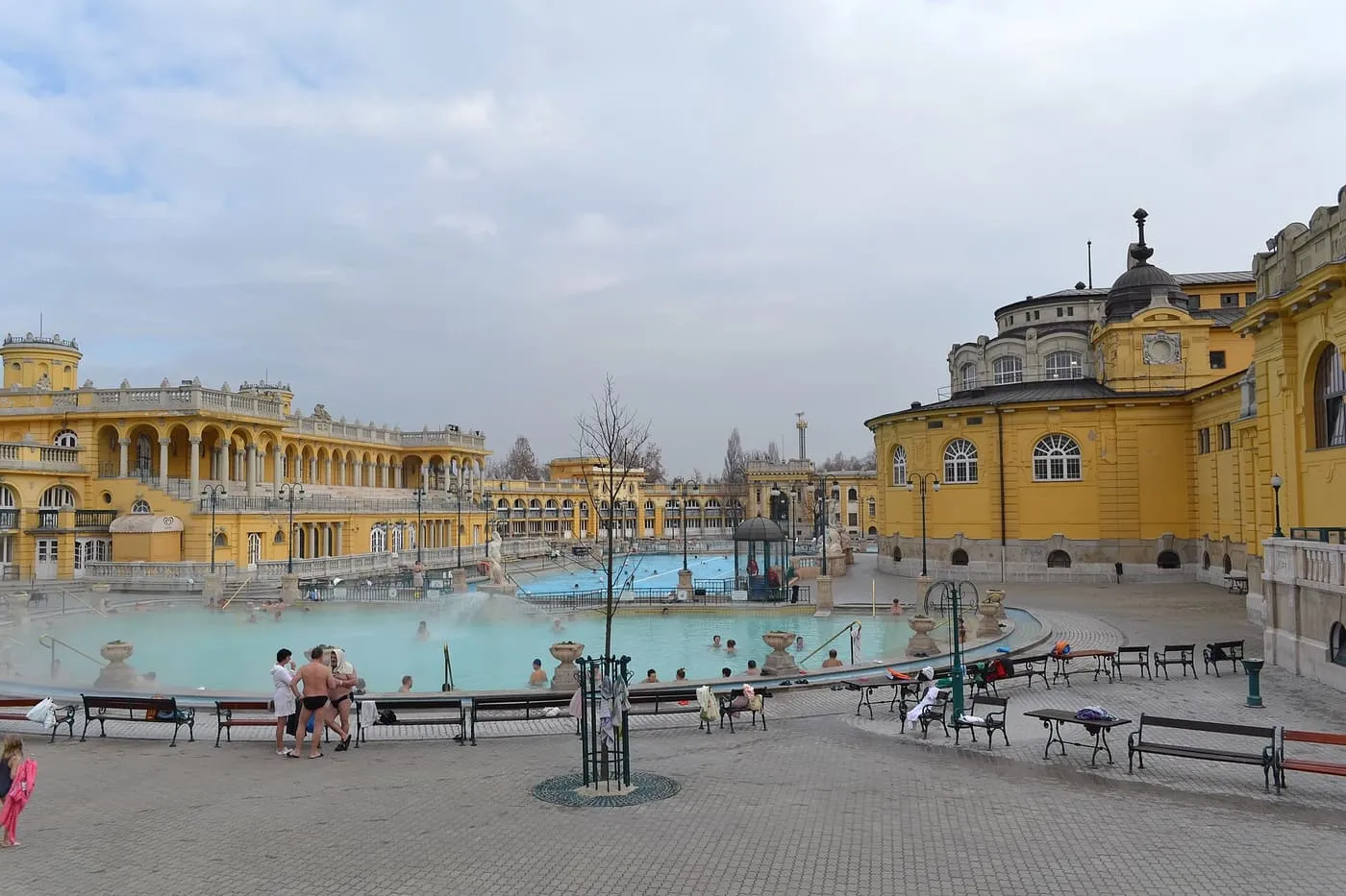 The outdoor thermal pools of the Széchenyi Baths, ringed by benches and the yellow bath buildings, with a few bathers in the water