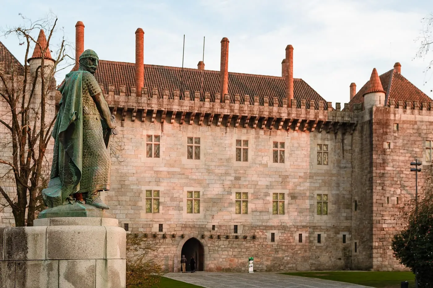 The stone front of the Paço dos Duques de Bragança, with leaded windows, a battlemented cornice and tall brick chimneys, and a bronze statue of a mail-clad knight in the foreground